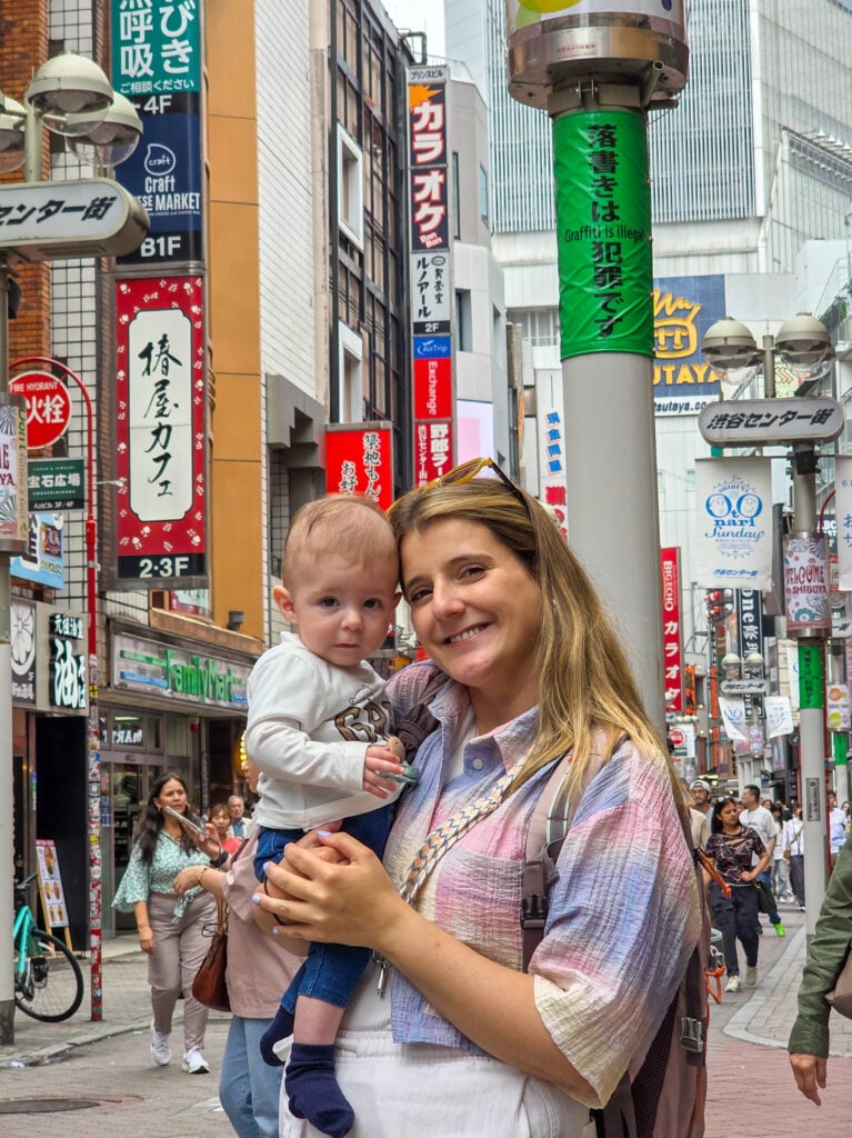 Mãe a segurar um bebé numa rua movimentada de Tóquio, rodeada por letreiros coloridos e edifícios típicos da cidade.