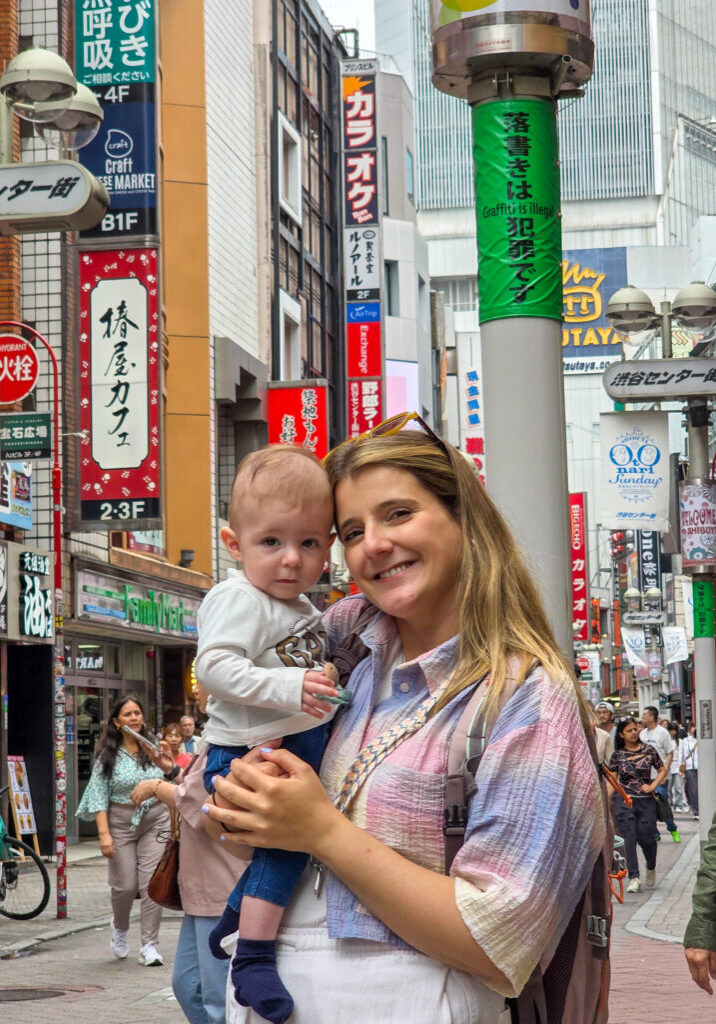 Mãe a segurar um bebé numa rua movimentada de Tóquio, rodeada por letreiros coloridos e edifícios típicos da cidade.