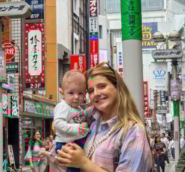 Mãe a segurar um bebé numa rua movimentada de Tóquio, rodeada por letreiros coloridos e edifícios típicos da cidade.